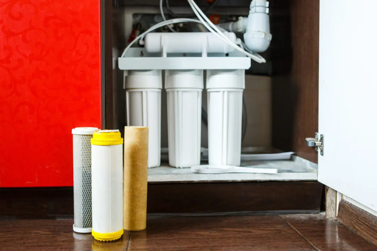 Water filtration system under a sink with three different filter cartridges displayed in front. The setting is a kitchen with a warm, organized feel.